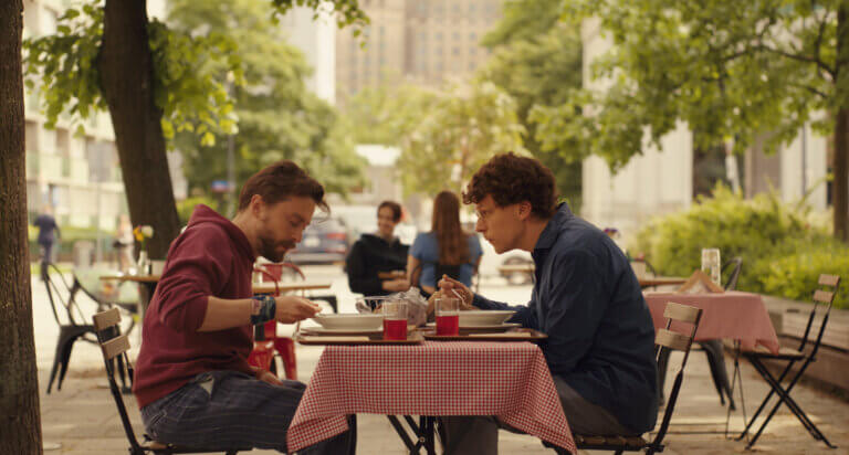 Kieran Culkin and Jesse Eisenberg eating lunch in a park in the film A REAL PAIN.